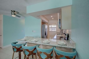 A modern kitchen with a marble countertop, four bar stools, a refrigerator, and a ceiling fan.