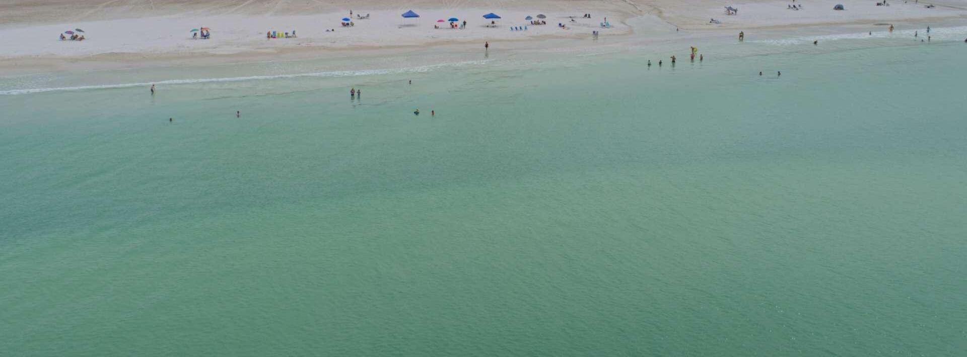 Une vue aérienne d’une plage avec une eau turquoise et limpide, un rivage sablonneux, ainsi que plusieurs parasols et des personnes.