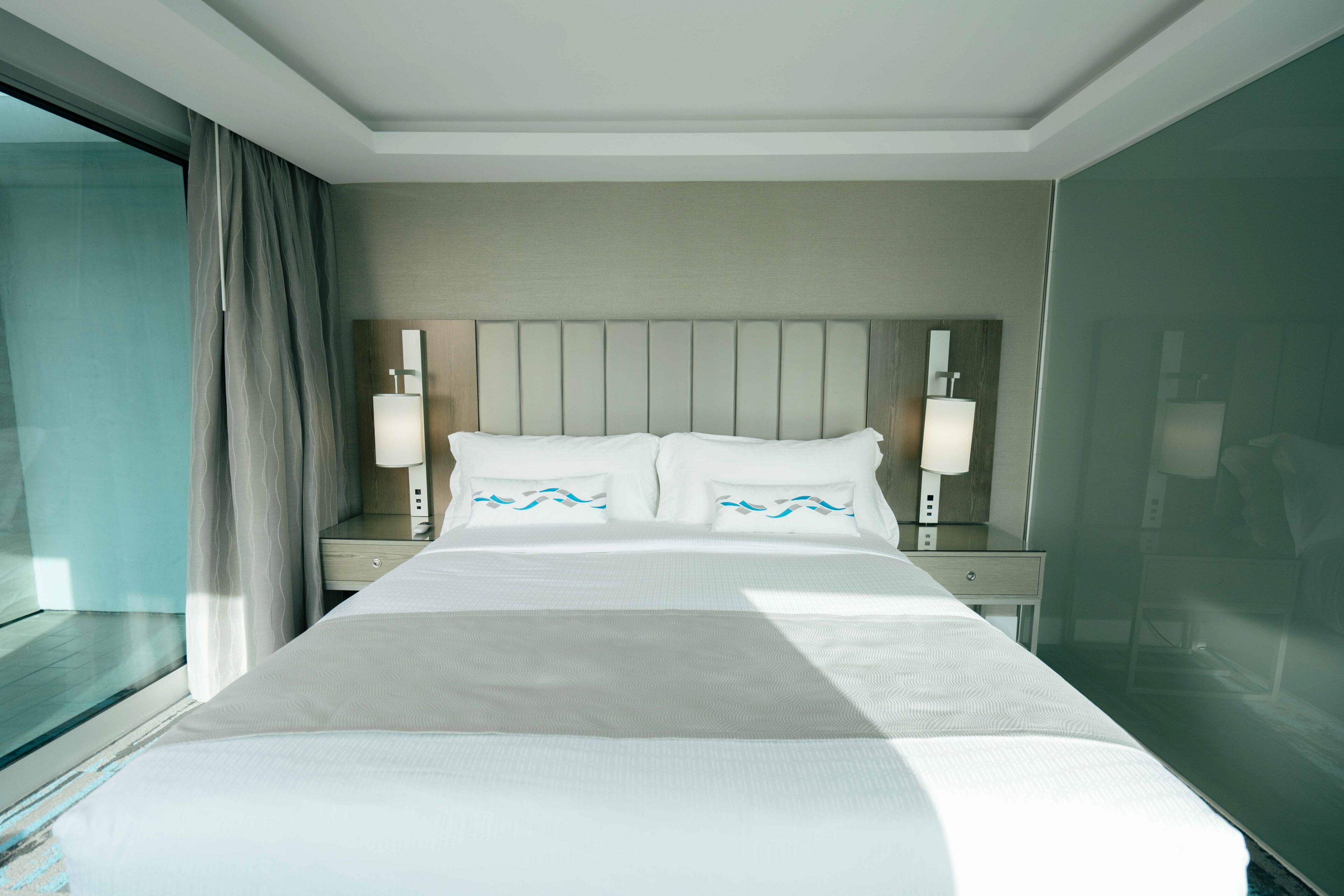 A hotel room with a large bed, two bedside tables with lamps, a headboard, and a view of the outside through a glass door.