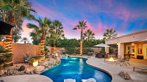 A swimming pool with a hot tub, surrounded by palm trees and a sunset sky.