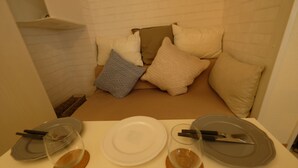 Ein Essbereich mit einem weißen Tisch, gedeckt für zwei Personen, inklusive Teller, Besteck und einem Glas. Der Raum verfügt über eine weiße Ziegelwand und beige Kissen auf einer niedrigen Couch.
