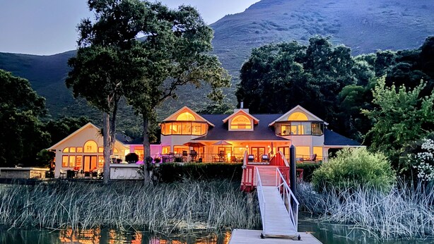 A lakeside property with a dock, a large house with multiple windows, and a mountain in the background.