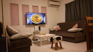 A living room with a brown sofa, a white coffee table, a small stool, and a television displaying a meal. - COCO Shinjuku (Tokyo)