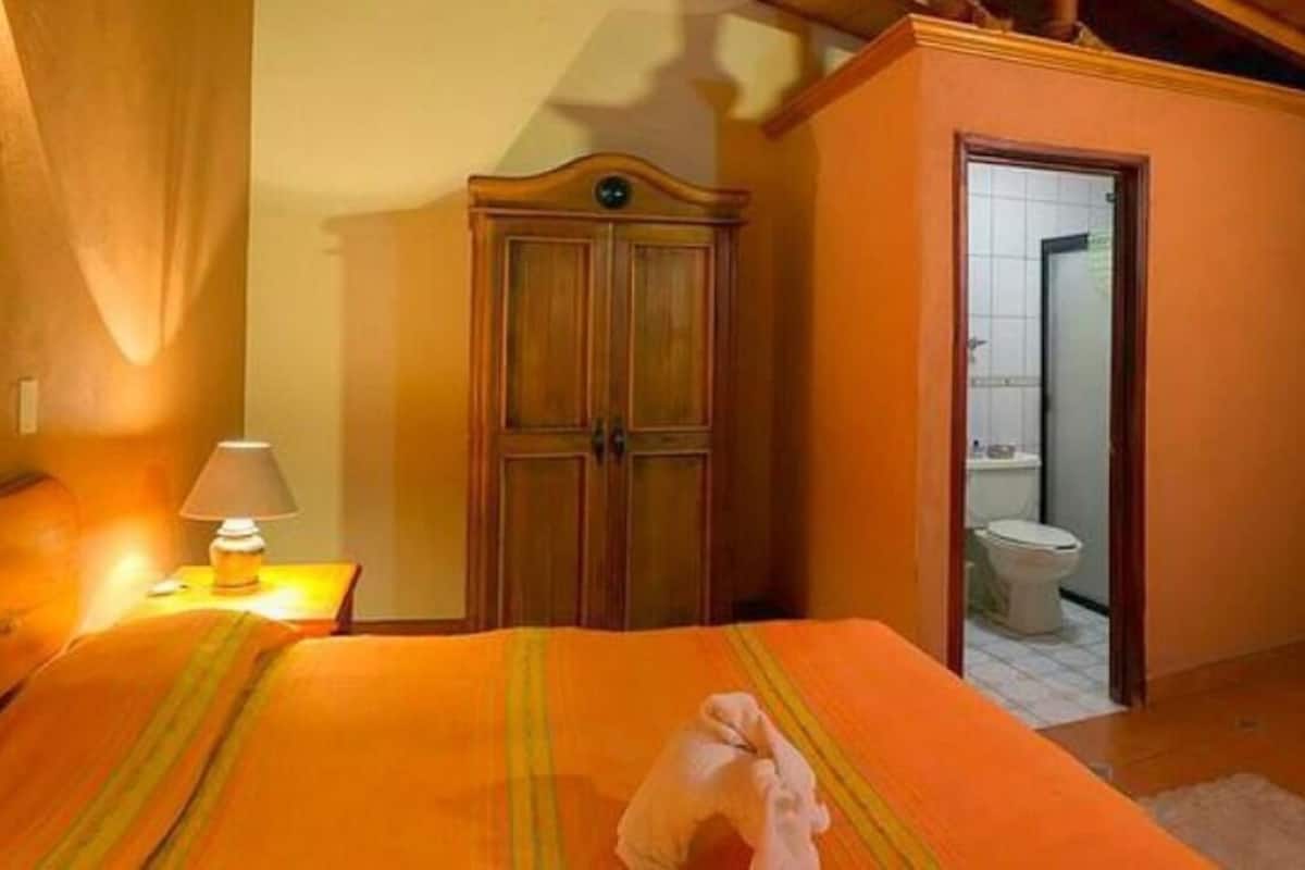 A hotel room with a bed, a wooden wardrobe, a bathroom visible through an open door, and a chair.