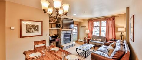 A cozy living room with a fireplace, a dining area with a wooden table set for four, and a comfortable seating area with a sofa and a TV.