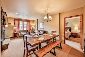 A dining area with a wooden table, benches, and a chandelier. There is a sofa and a fireplace in the background.