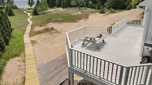 A wooden deck with a picnic table, overlooking a beach and forested area. - Stunning Lakefront 5BR 5000 sq ft Dune Alright (Baileys Harbor)