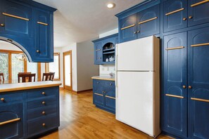 A kitchen with blue cabinets, a white refrigerator, wooden flooring, and a window with a view.