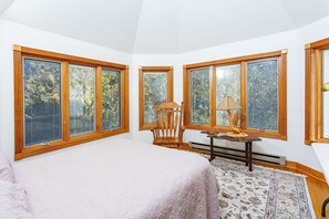 A bedroom with a large bed, a wooden chair, a small table with a lamp, and a view of trees through large windows.