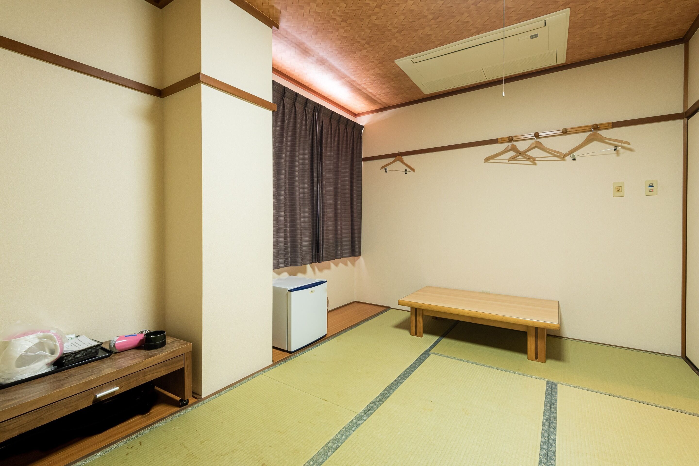 Una habitación con suelo de tatami, un banco de madera, una pequeña mesa con un objeto rosa, un electrodoméstico blanco y un perchero.