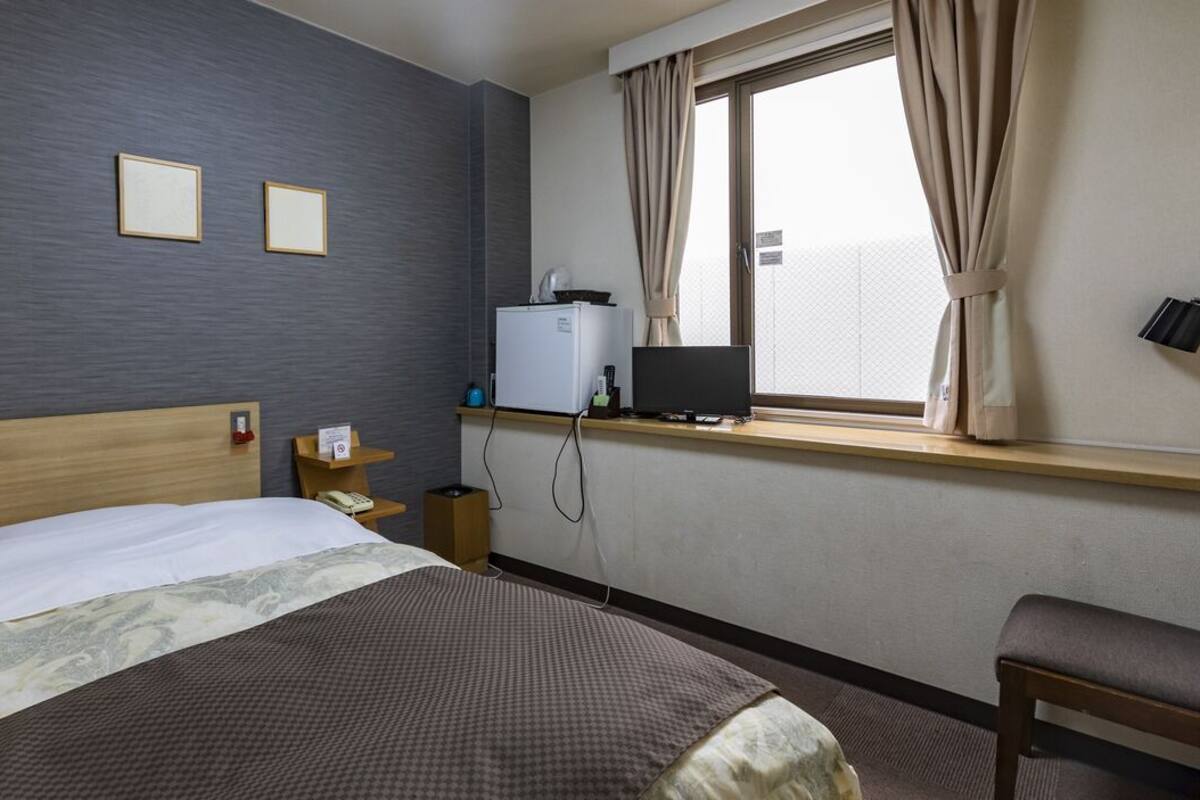 A hotel room with a bed, a desk with a computer, a small refrigerator, and a window with curtains.