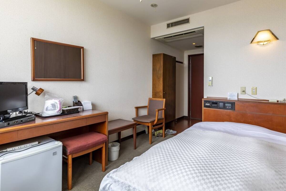 A hotel room with a bed, desk, chair, and a small refrigerator.