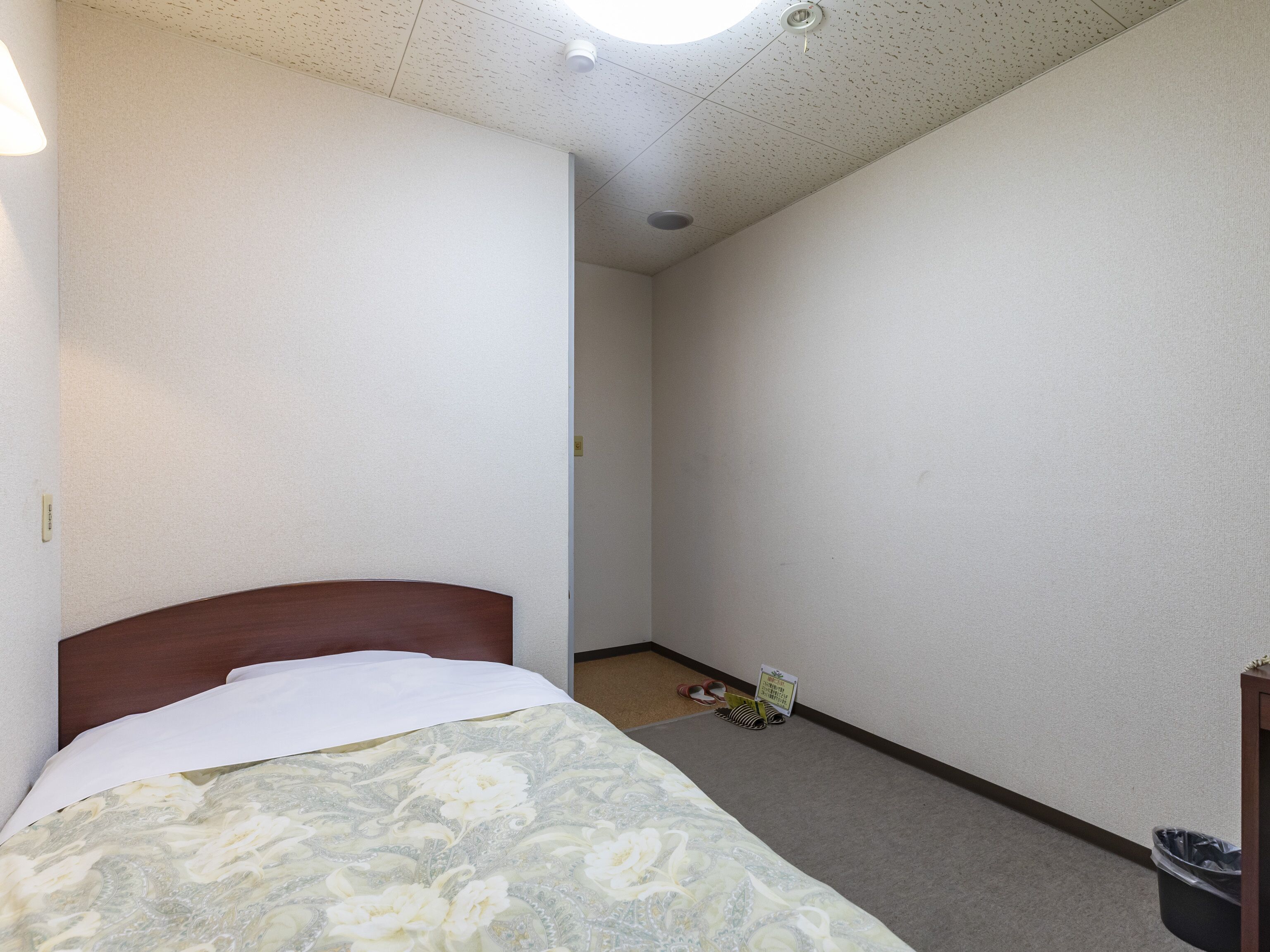 Una habitación pequeña y escasamente amoblada con una cama individual, un cubo de basura pequeño y un par de zapatos en el suelo.