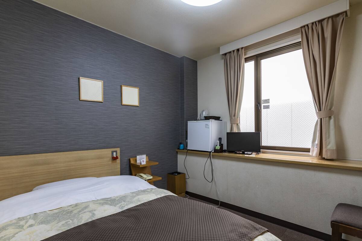 A hotel room with a bed, a desk with a computer, a window with curtains, and a wall-mounted TV.