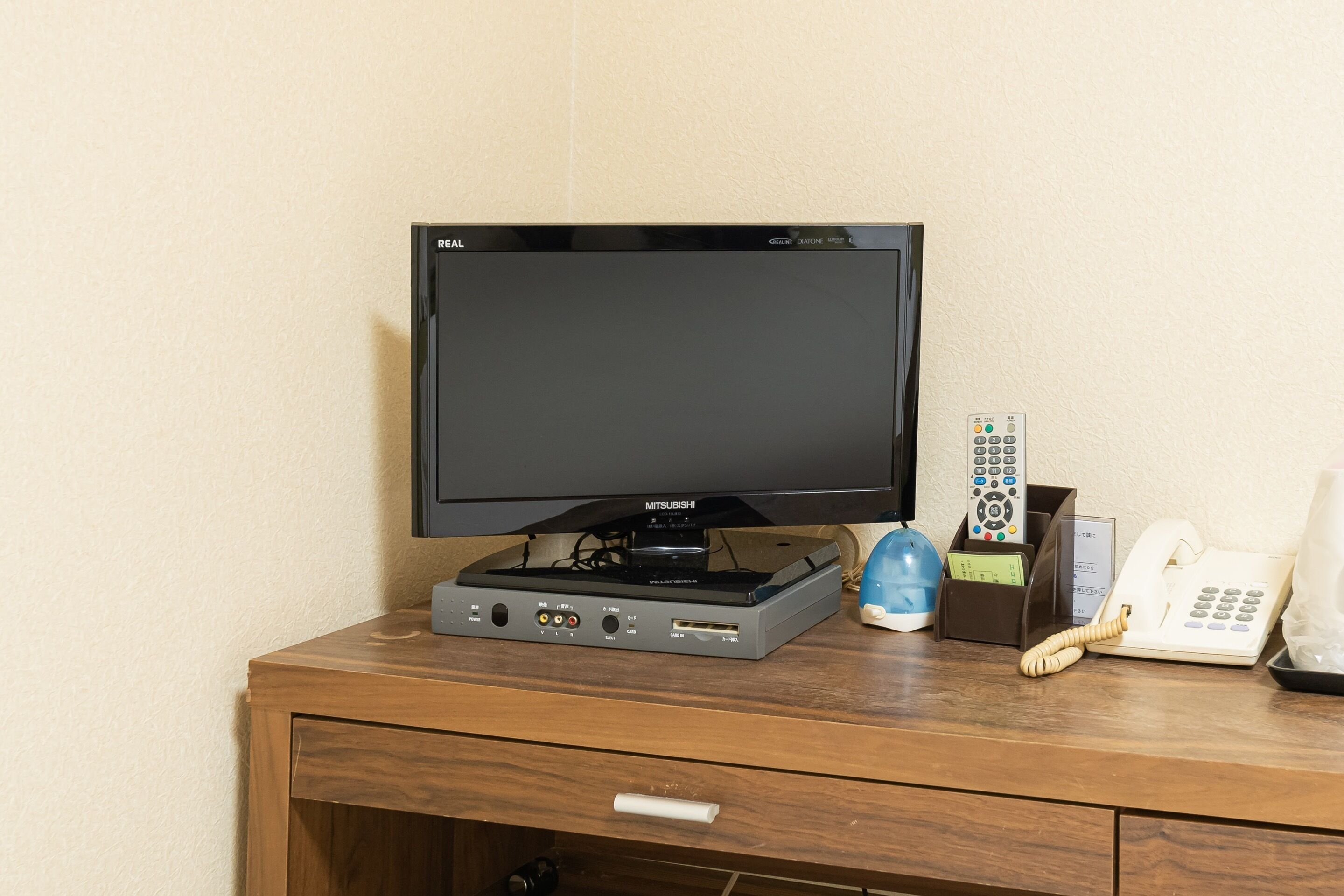 A television on a wooden desk with a remote control, a telephone, and a small blue container.