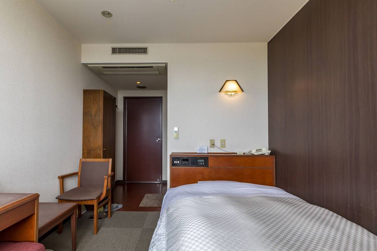A hotel room with a bed, a desk, a chair, a dresser, and a wall-mounted lamp.