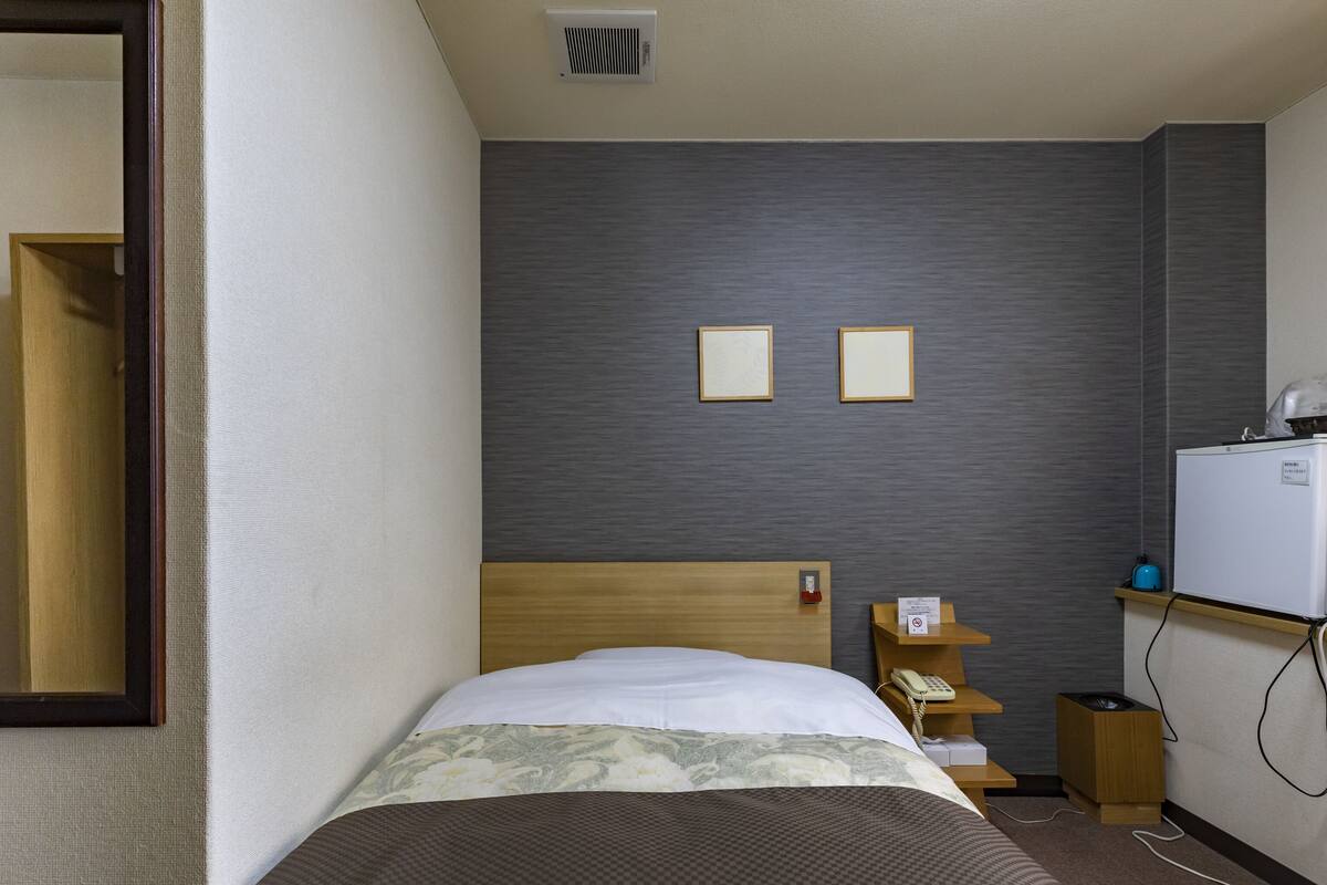 A hotel room with a bed, a wooden headboard, a small bedside table with a phone and a sign, a wall-mounted heater, and a refrigerator.