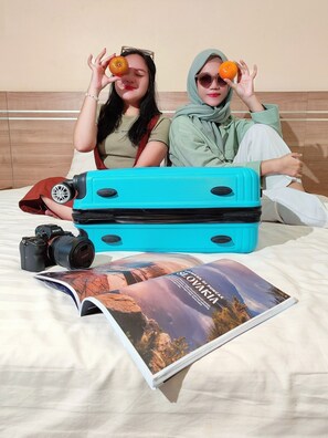 Two individuals sitting on a bed with a blue suitcase, a camera, and a travel magazine. - Gapura Residence By Sinergi (Semarang)