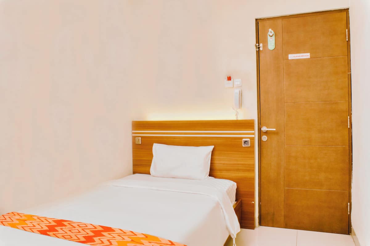 A hotel room with a wooden headboard, a white bed, a patterned throw, a door with a sign, and a wall-mounted phone.