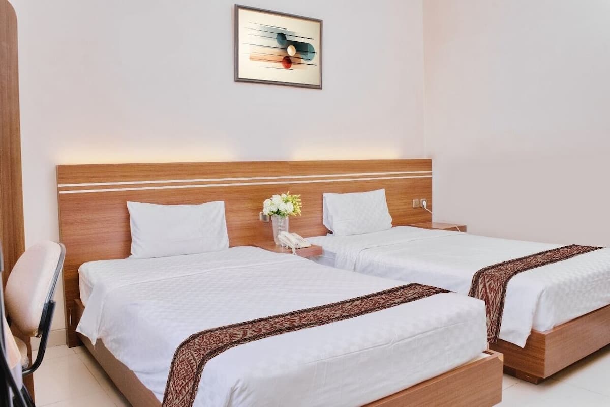 A hotel room with two single beds, a wooden headboard, white bedding, a bedside table with a vase of flowers, and a framed abstract artwork on the wall.