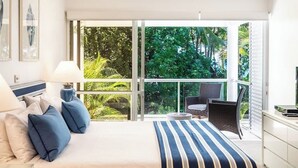 A bedroom with a large bed, a nightstand, a lamp, a TV, and a balcony with a view of greenery.