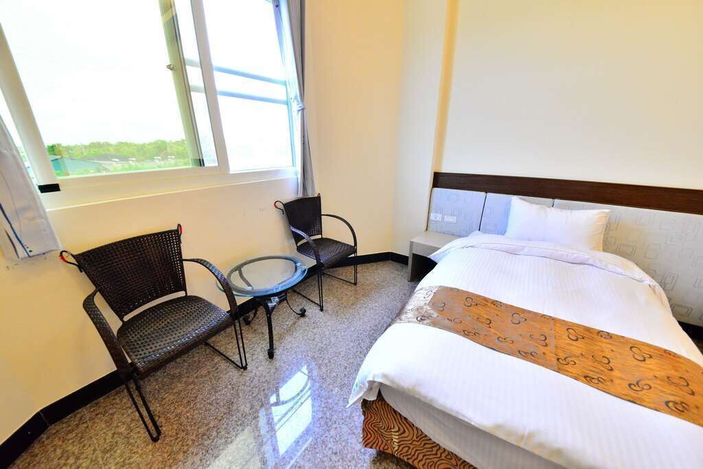 A hotel room with a bed, a small table, two chairs, a window, and a nightstand.