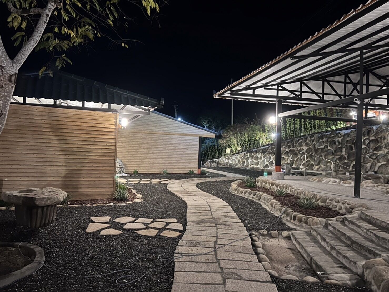 A well-lit outdoor area with a stone pathway leading to a covered seating area, surrounded by a landscaped garden with a stone wall and a wooden building.