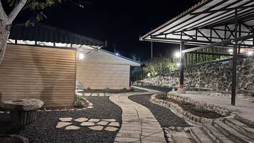A well-lit outdoor area with a stone pathway leading to a covered seating area, surrounded by a landscaped garden with a stone wall and a wooden building.