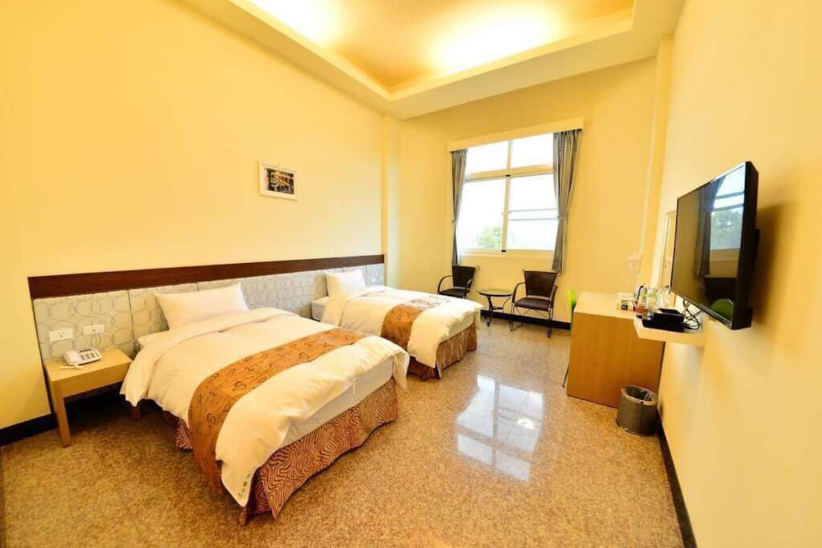 Habitación de hotel con dos camas, un escritorio, una silla, un televisor y un ventanal con cortinas.