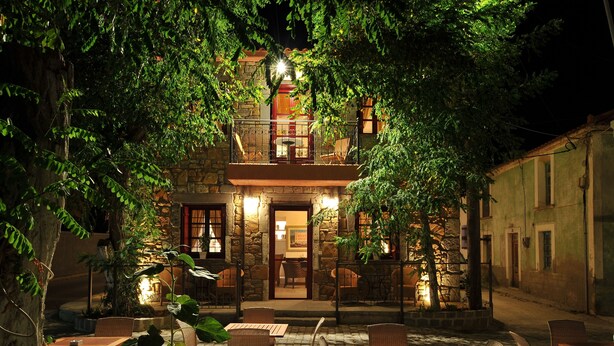 An outdoor patio area with stone walls, wooden tables, wicker chairs, and a stone building with a balcony in the background. - Lively Lofts by The Varos Residences (Lemnos)