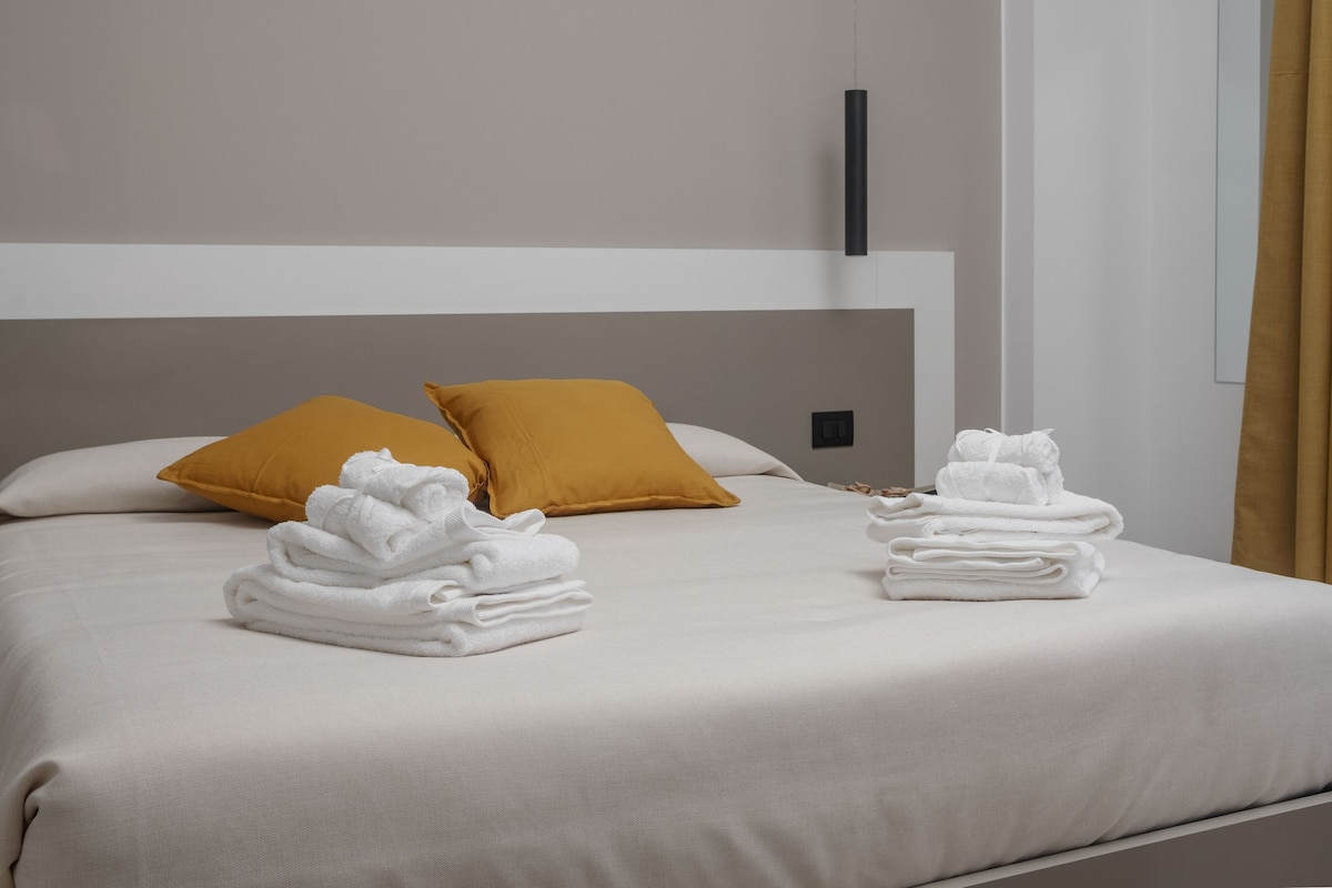A neatly made bed with white linens and two mustard-colored pillows, a stack of white towels, and a simple, modern room design.