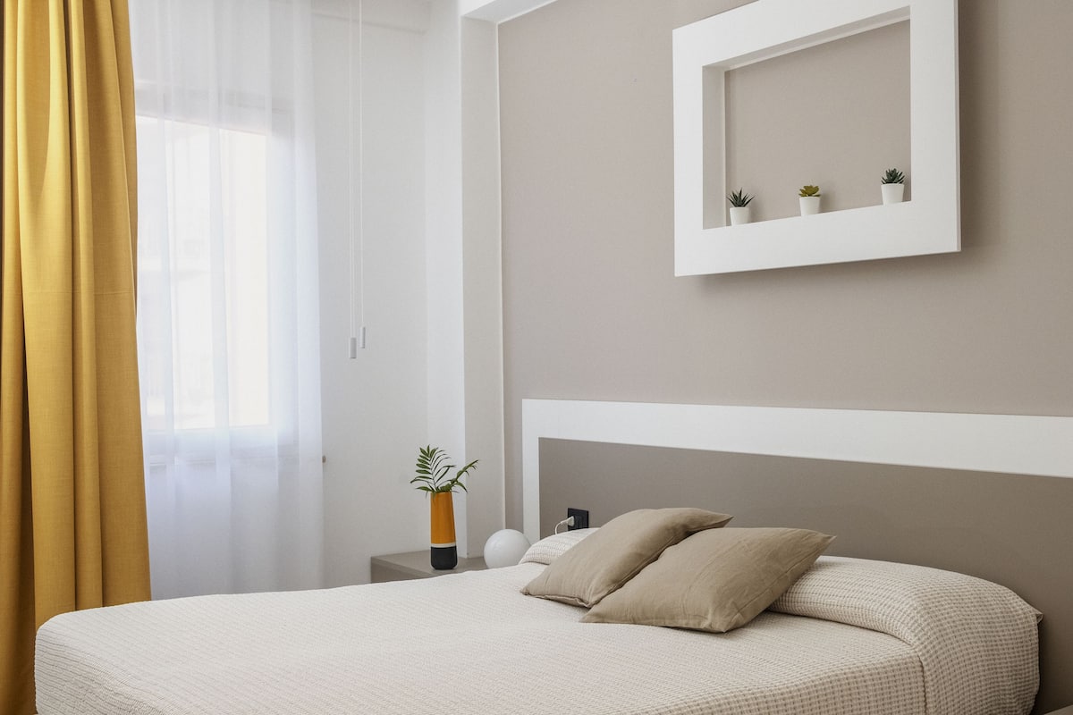 A modern bedroom with a large bed, a bedside table with a vase, and a wall-mounted shelf with small plants.