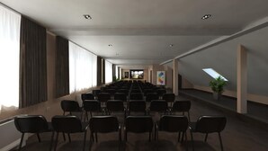 Un salón de conferencias con filas de sillas, ventanas amplias y una planta a la derecha.
