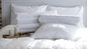 Una cama bien hecha con ropa de cama y almohadas blancas, una bandeja de madera con libros y un objeto verde, y un muro de ladrillo al fondo.