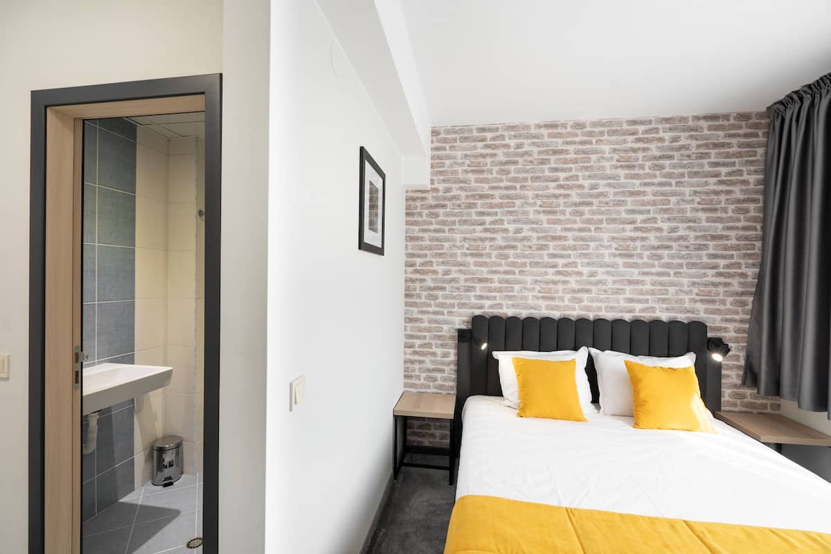 A hotel room with a brick-patterned wall, a bed with white and yellow bedding, a bedside table, and a small trash bin.