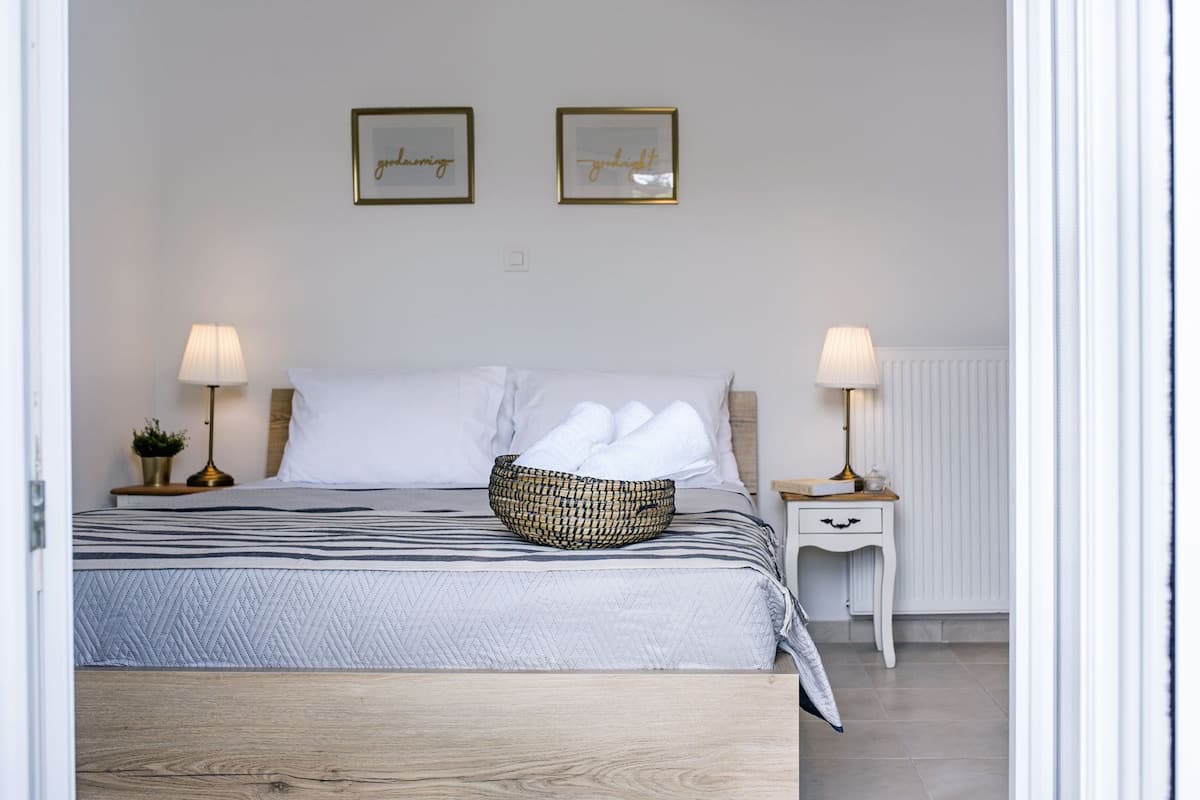 A neatly made bed with white linens, a striped blanket, and a woven basket on the bed. There are two bedside lamps, a small plant, and a white nightstand with a drawer. Two framed pictures hang on the wall above the bed.