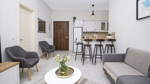 Espai habitable petit amb un sofà, dues butaques, taula rodona petita amb un vas, zona de cuina amb nevera i taula de menjador amb tamborets.