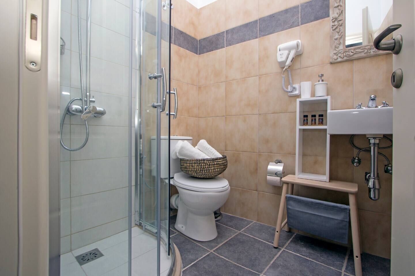 A bathroom with a glass shower enclosure, a toilet with a basket of towels, a sink with a mirror, and a hairdryer on the wall.