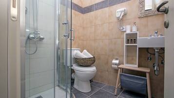 A bathroom with a glass shower enclosure, a toilet with a basket of towels, a sink with a mirror, and a hairdryer on the wall.