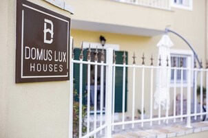Segnaletica per Domus Lux Houses, con recinzione bianca e porta verde sullo sfondo.