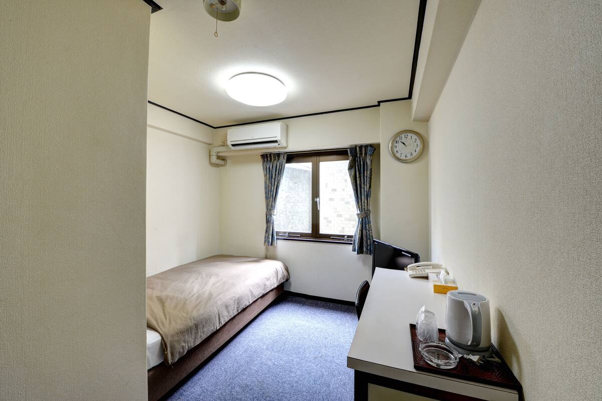A small, single-bed room with a desk, a clock, an air conditioner, and a window with curtains.