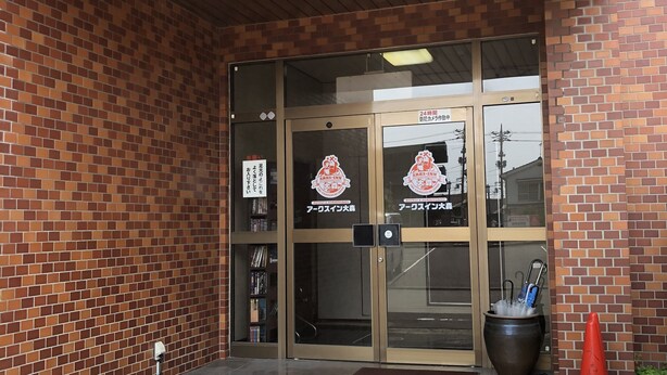 A hotel entrance with a brick facade, a glass door, and a small outdoor seating area with a potted plant and a red cone. - Arks Inn Omori (Ube)