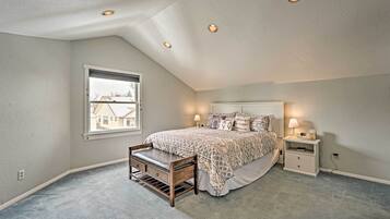 Une chambre avec un plafond en pente, un grand lit avec une literie à motifs, une table de chevet avec une lampe et une fenêtre donnant sur des immeubles.