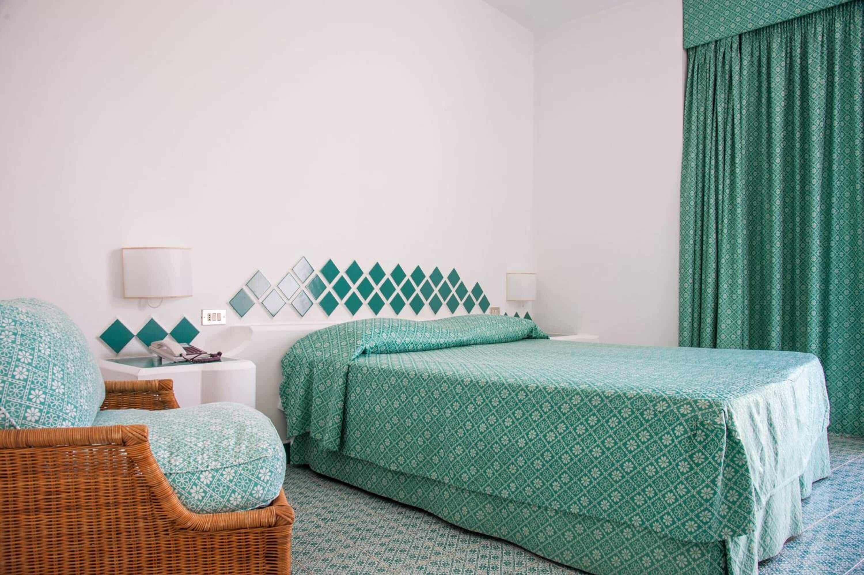 Una habitación de hotel con una cama, dos mesitas de noche, una silla de mimbre y un adorno de pared con estampado.