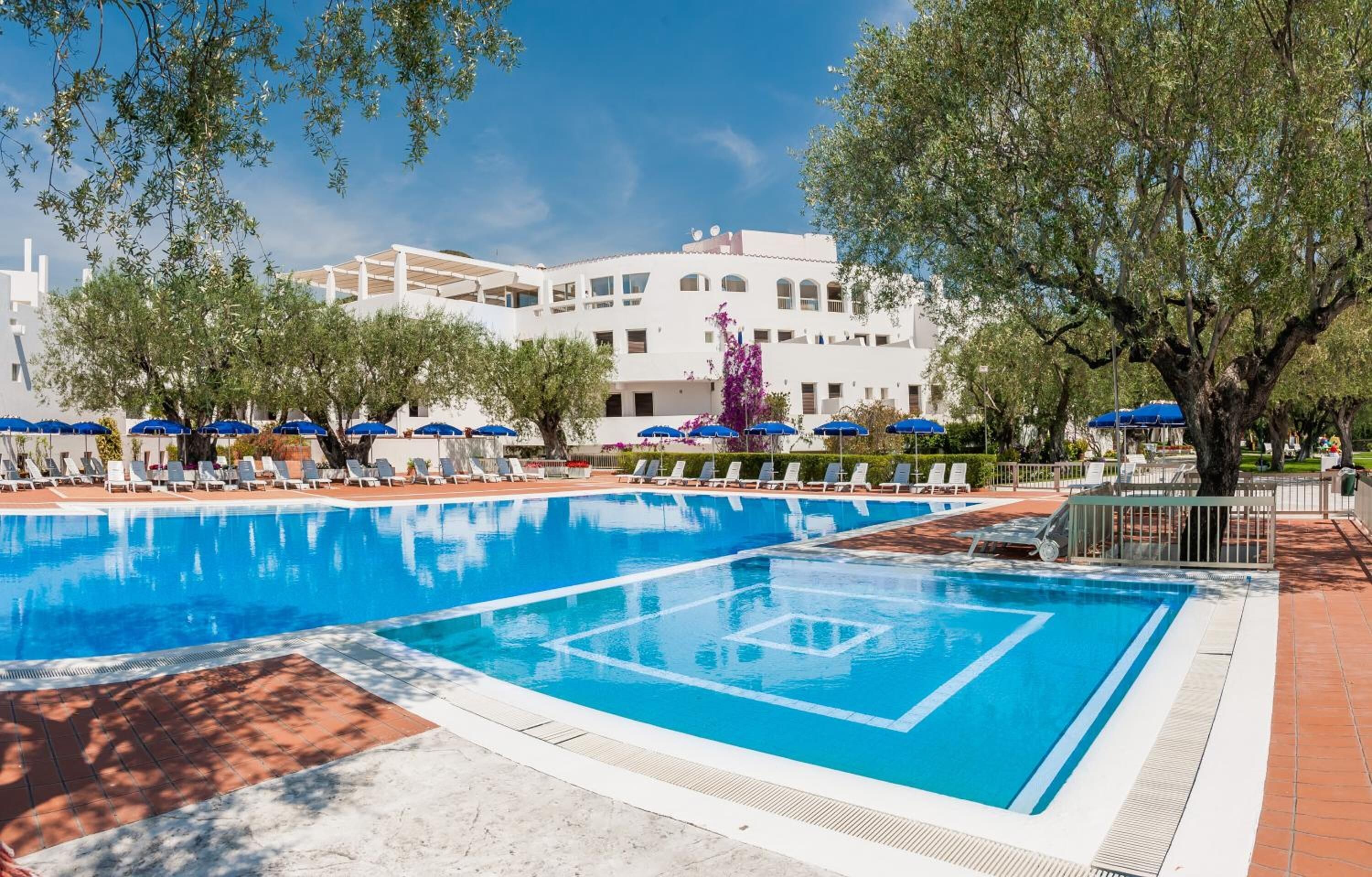 Una piscina con lettini e ombrelloni, immersa nel verde e affiancata da un edificio con facciata bianca.