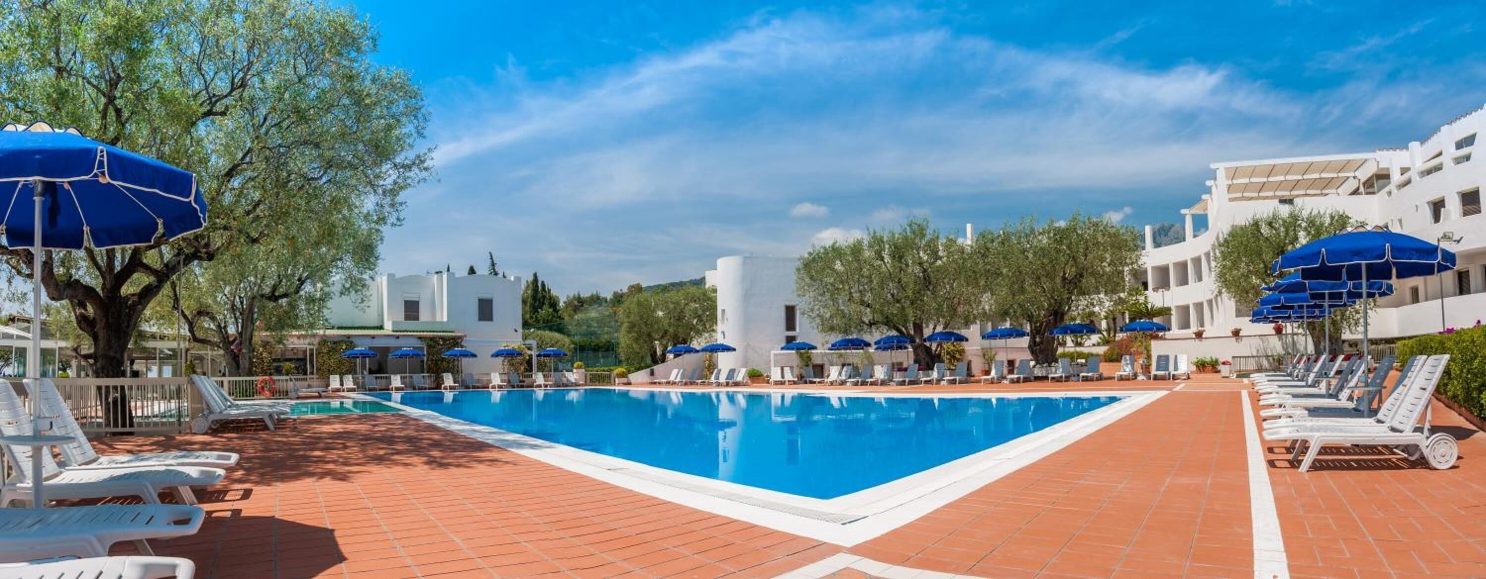 Ein Pool mit Liegestühlen und Sonnenschirmen, umgeben von Bäumen und weißen Gebäuden.