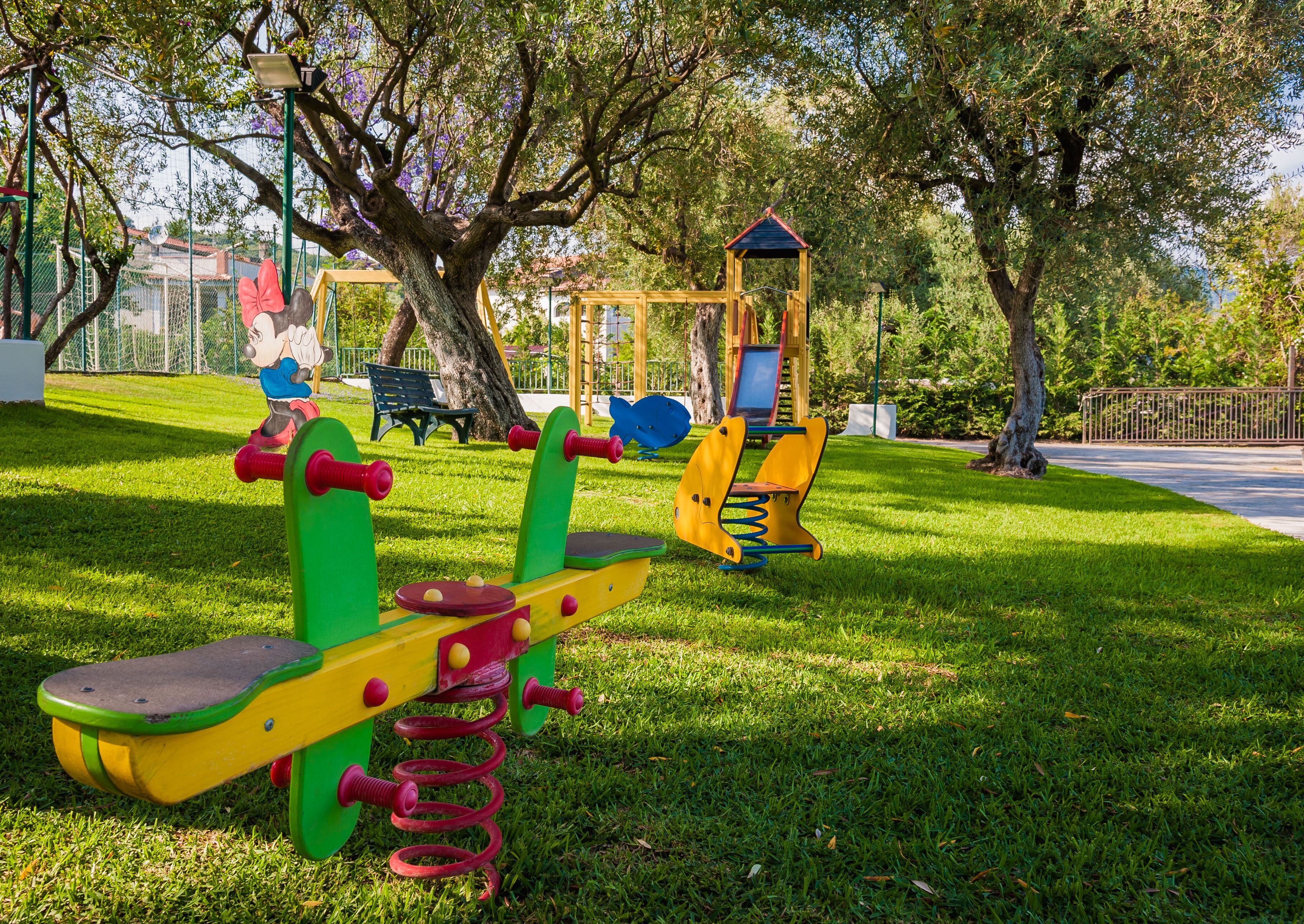 A playground with various play structures, including a seesaw and a climbing frame, surrounded by grass and trees.