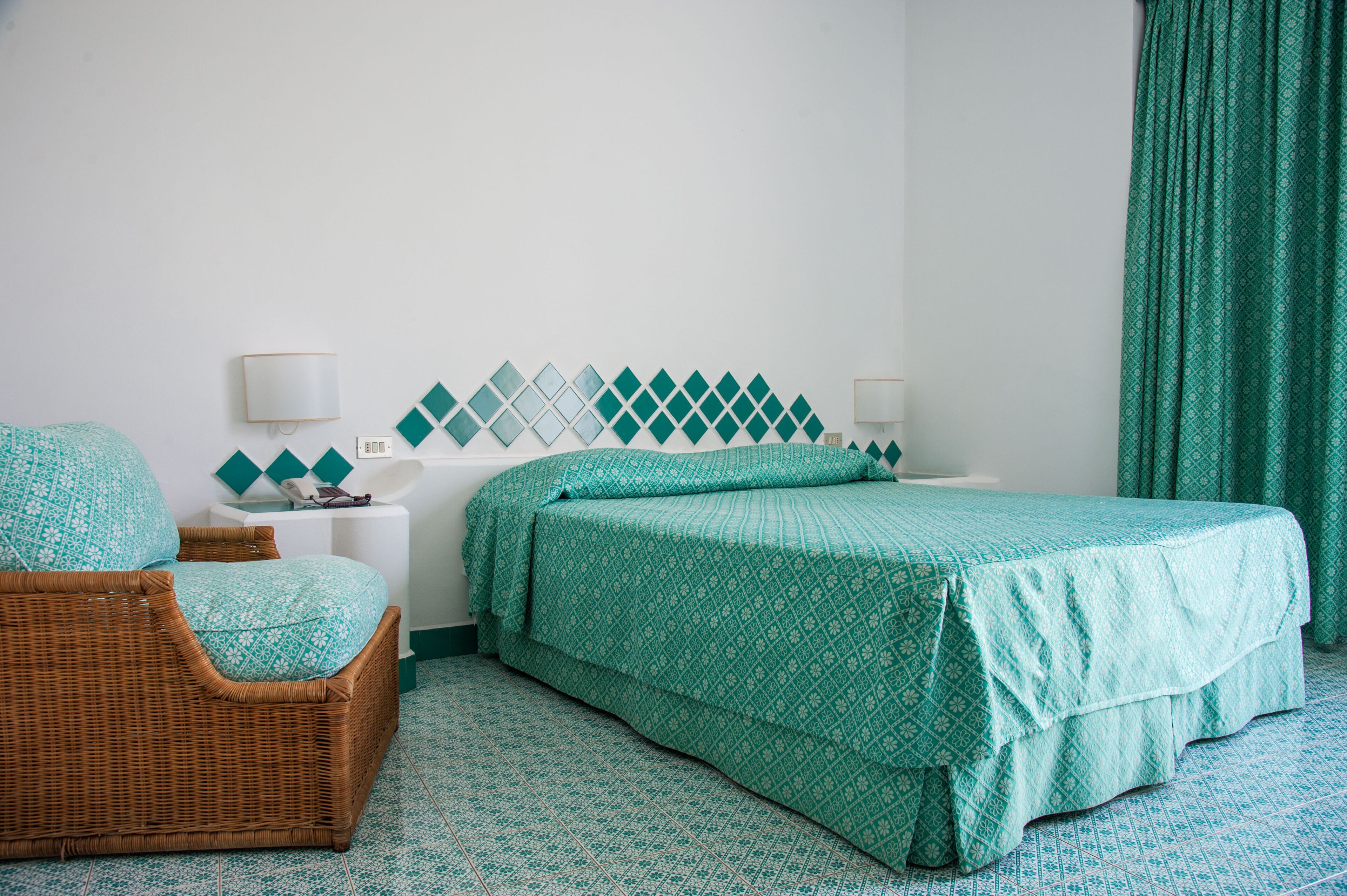 A hotel room with a bed, a wicker chair, a nightstand, and a wall with a geometric pattern.