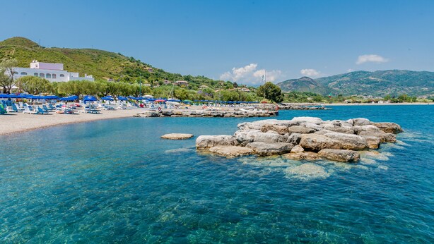 A beach with clear blue water, rocky formations, and a row of beach umbrellas. - Hotel Torre Oliva (Santa Marina)