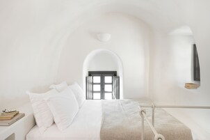 Un dormitorio minimalista con cama, almohadas, una mesita de noche con libros y una puerta que da a otra habitación.